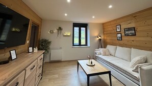 A modern living room with a wooden floor, a sofa, a coffee table, a TV, and a window with a view of greenery.