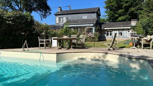 A swimming pool with a wooden deck and lounge chairs, surrounded by a garden and a house with a grey roof.