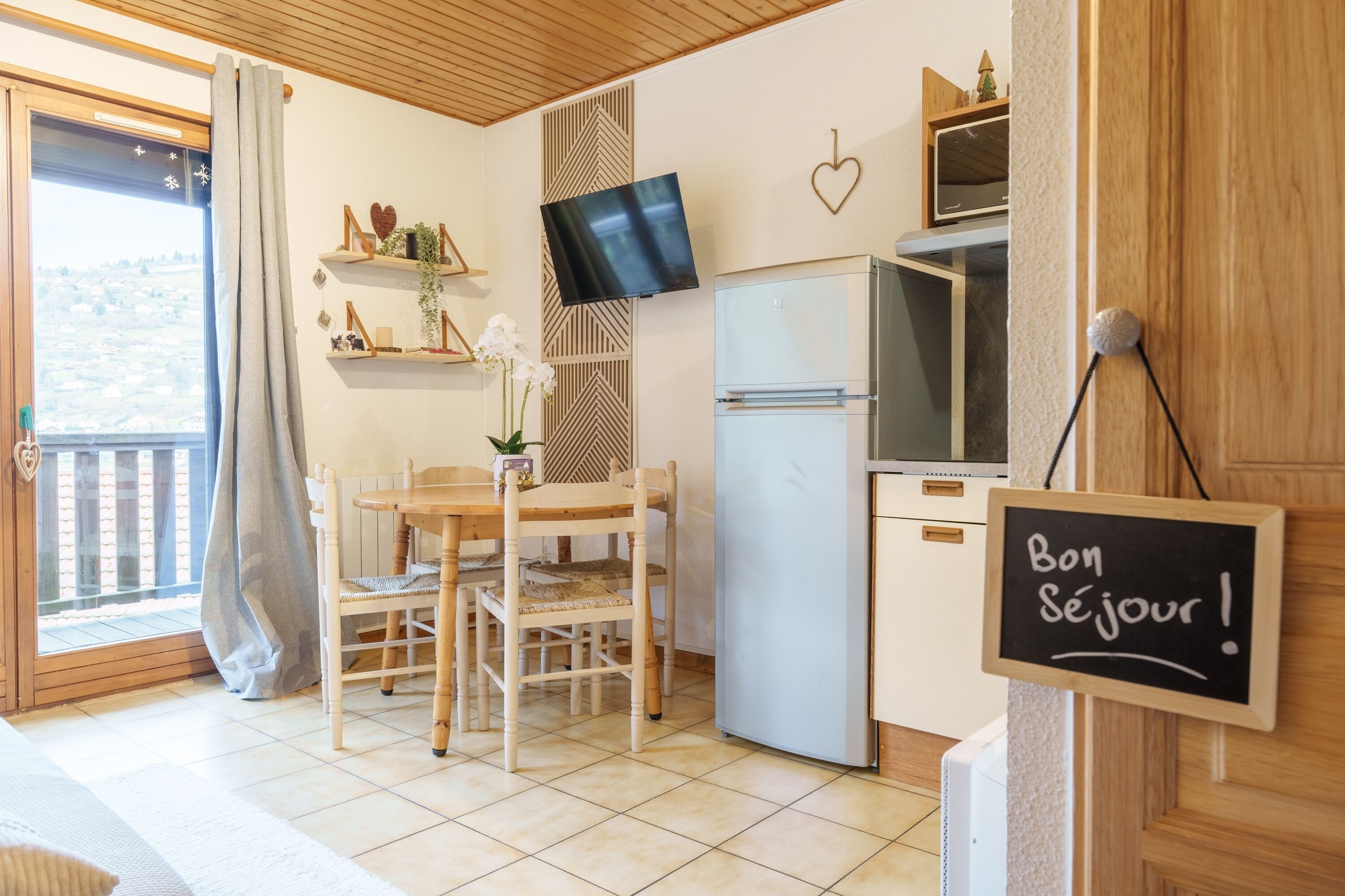 A compact kitchen with a dining table, chairs, a refrigerator, a TV, and a sign saying "Bon Séjour!".