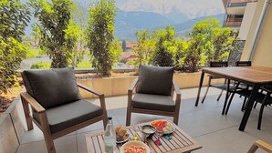 Een terras met twee comfortabele stoelen, een houten tafel met eten en drinken, en uitzicht op bergen en groen.