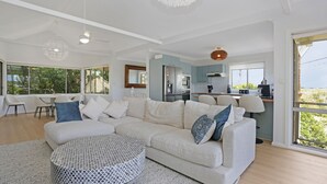 A spacious living room with a large L-shaped sofa, a round coffee table, and a dining area with a table and chairs. A kitchen is visible in the background with modern appliances.