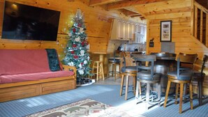 A cozy cabin interior with a wooden interior, a dining area with metal chairs, a Christmas tree, and a sofa with a red blanket.