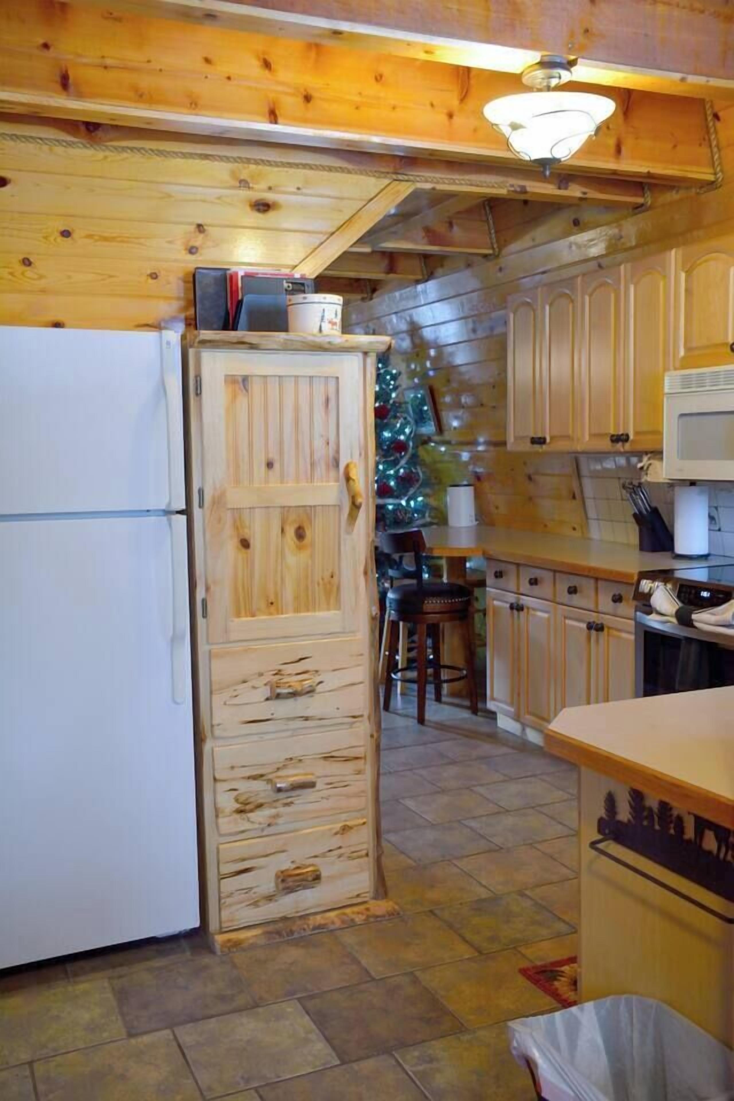 Una cuina compacta amb armaris de fusta, un refrigerador blanc i un microones. Es veu un petit armari de fusta amb calaixos, i un arbre de Nadal al fons.