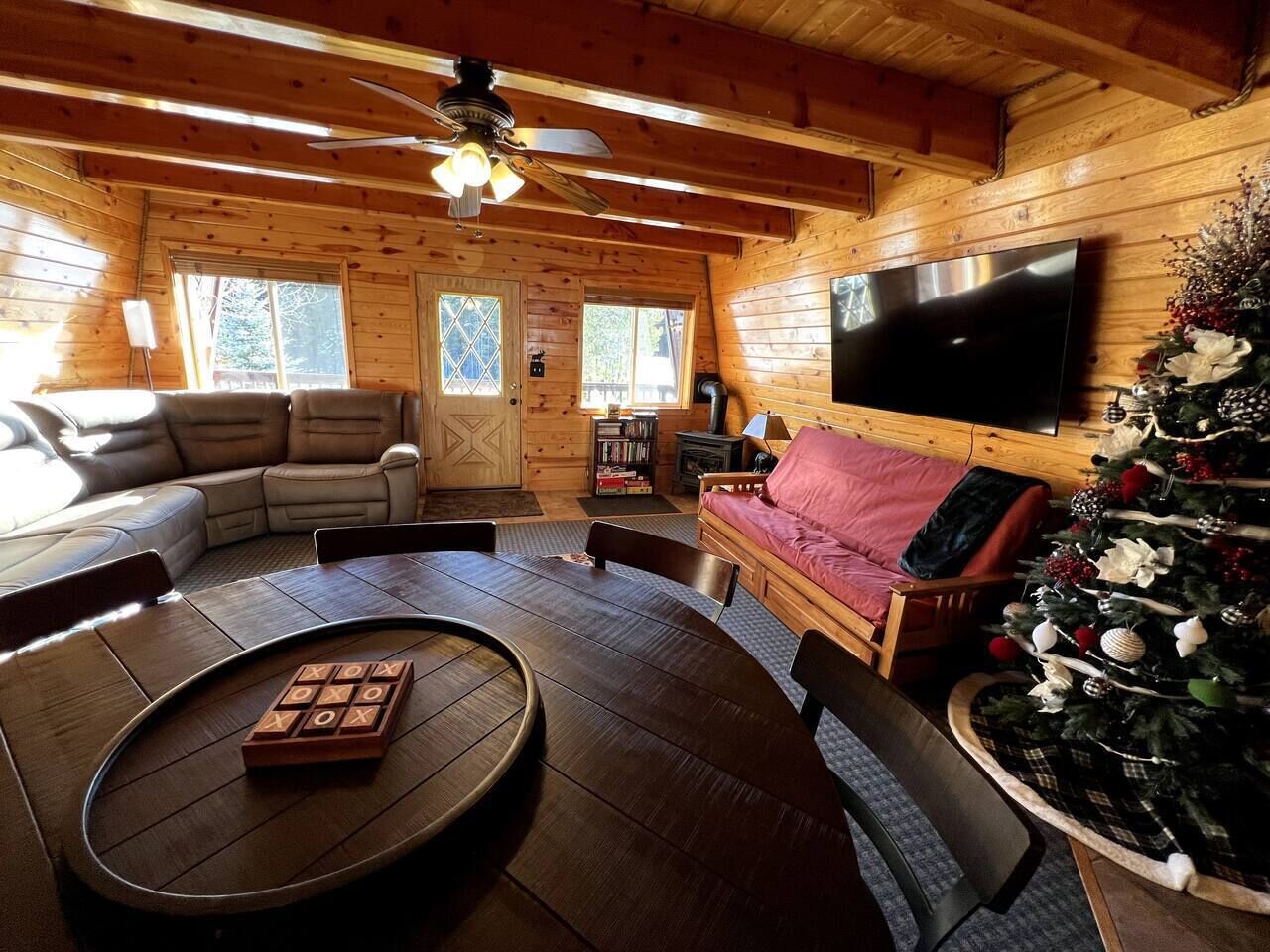 A cozy cabin interior with a wooden interior, a round dining table, a sofa, a television, and a decorated Christmas tree.