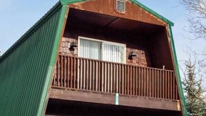 A two-story cabin with a green exterior, a balcony on the upper floor, and a stone foundation.