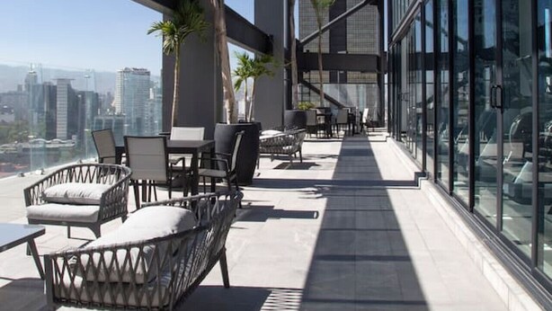 A rooftop terrace with outdoor seating, palm trees, and a city skyline view.