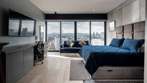 A modern bedroom with a large bed, a sofa, and a desk. It features a city view through large windows.
