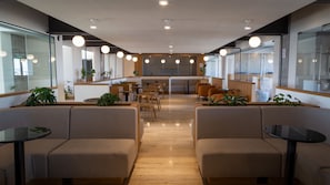 Relax in a modern space with cushioned benches and tables, surrounded by potted plants and large windows.