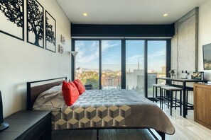A modern bedroom with a large bed, a desk with a chair, and a city view.