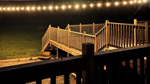 Una terraza de madera con barandilla, un área ajardinada y luces colgantes al fondo.