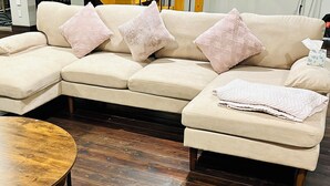 A beige sectional sofa with three pink cushions, a wooden coffee table, and a pair of yellow slippers on a wooden floor.