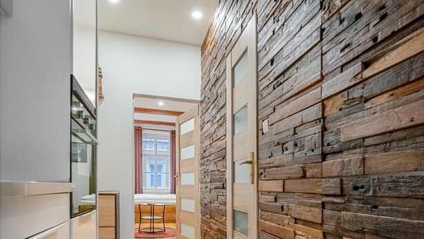 A hallway with a tiled floor, a wooden wall with glass panels, and a door leading to a room with a window and a table.