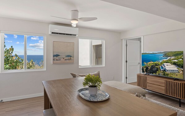 A modern living room with a dining table, a ceiling fan, a potted plant, and a television displaying a scenic view. - Beautiful SeaCrown Estate-Oceanfront- 6BR 5.5BA with Pool (St Thomas)