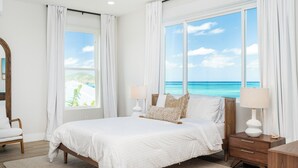 A bedroom with a bed, two bedside tables, a nightstand, a chair, and large windows with a view of the ocean. - Beautiful SeaCrown Estate-Oceanfront- 6BR 5.5BA with Pool (St Thomas)