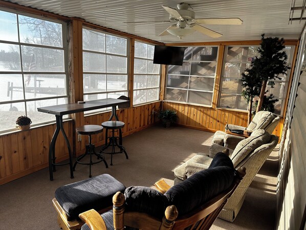 A cozy room with a wooden interior, a ceiling fan, a TV mounted on the wall, and a large window offering a view of the outdoors. - Charming 2-bedroom house with WiFi, AC on Knife Lake in Mora Minnesota. (Mora)