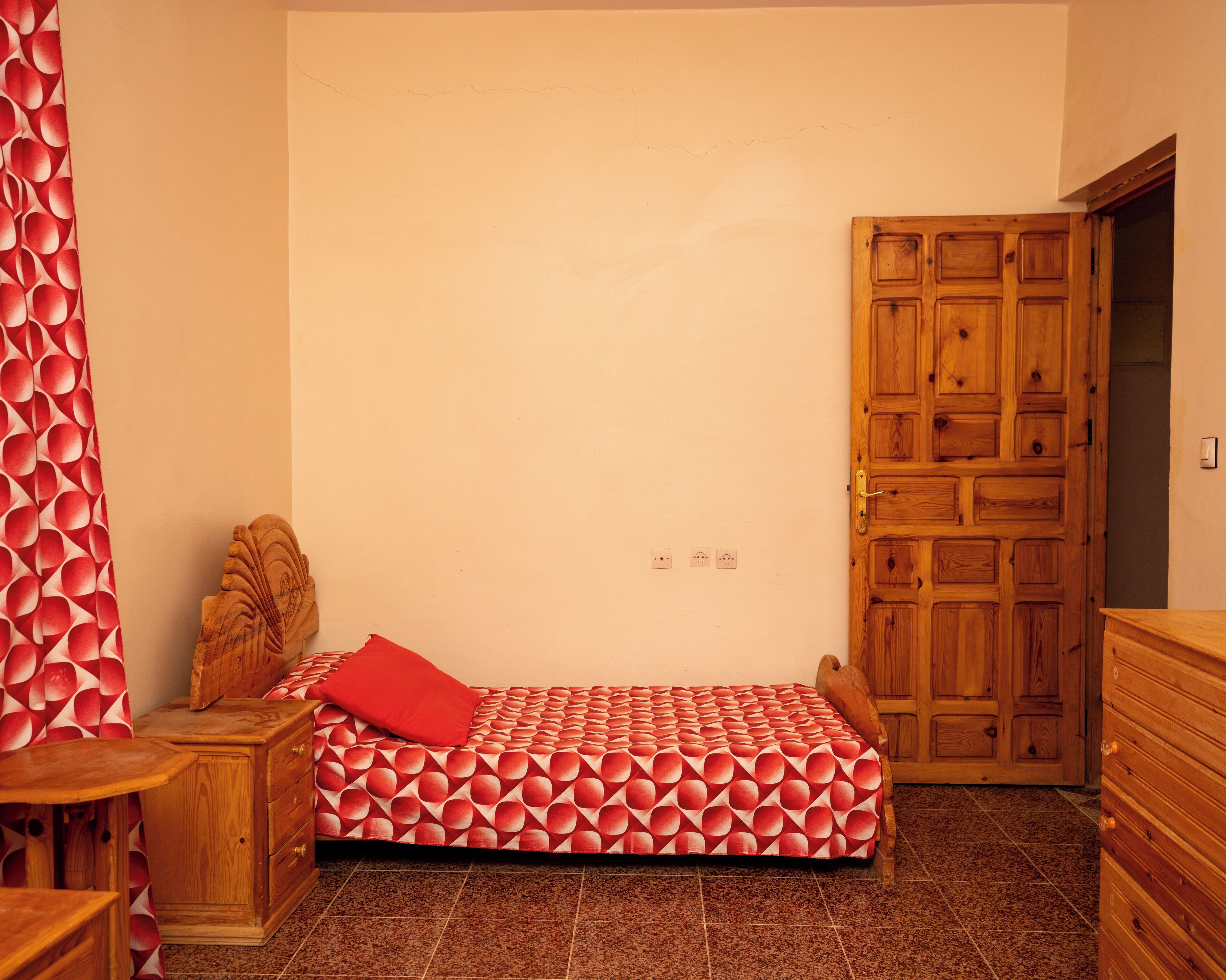 Ein Einzelbett mit geometrischem Muster auf der Bettdecke, ein Nachttisch aus Holz, ein rotes Kopfkissen und eine Holzkommode sowie eine Holztür.