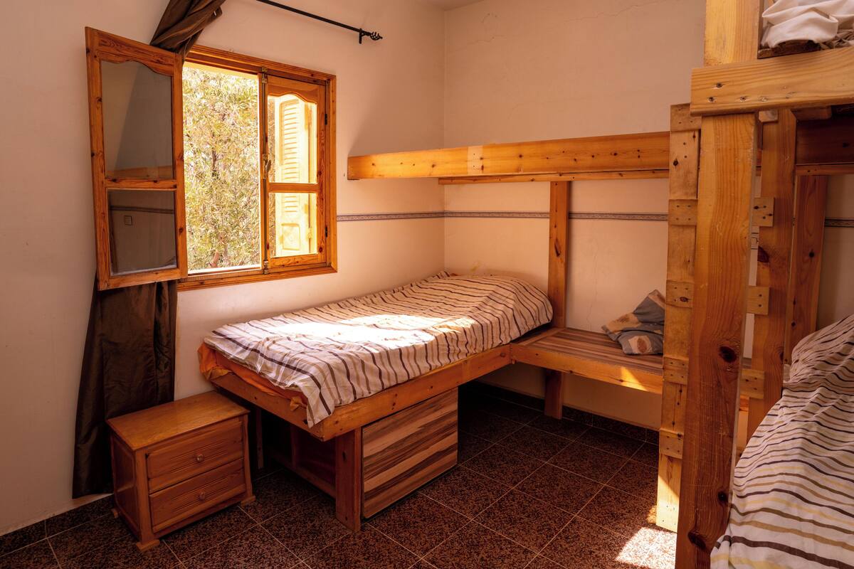 A small, single-room accommodation with a bunk bed, a wooden dresser, a bedside table, and a window with curtains.