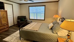 A bedroom with a bed, a ceiling fan, a window with blinds, a nightstand with a lamp, and a wooden dresser.