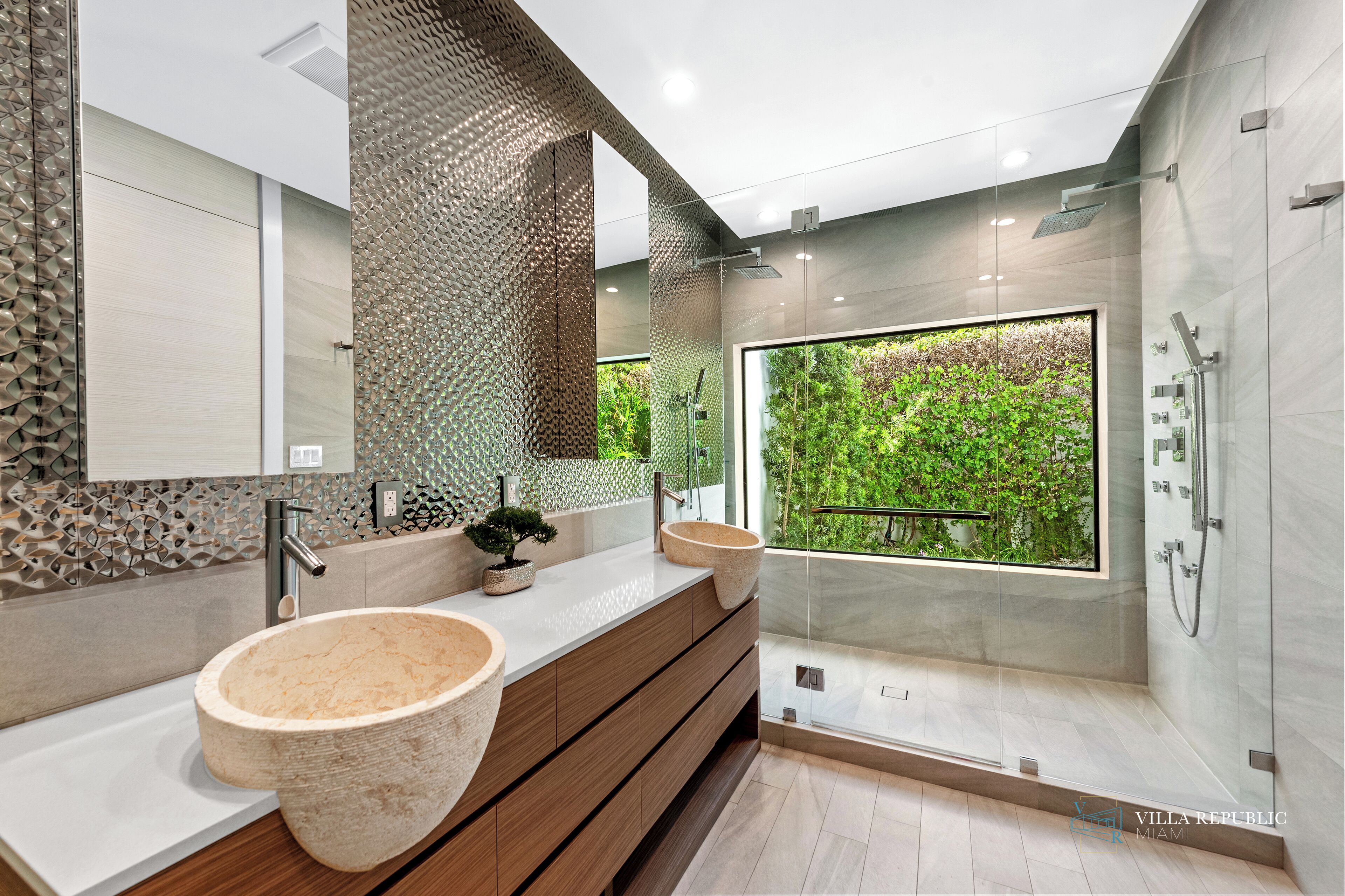 Un baño moderno con dos lavamanos grandes de piedra, un mueble de tocador de madera, una cabina de ducha de vidrio y vistas a la vegetación a través de un ventanal amplio.