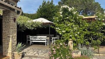 A stone patio with a white table and chairs under a pergola, surrounded by potted plants and flowering shrubs.
