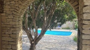 A stone archway with a tree in the center, leading to a pool area with stone flooring.