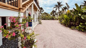 A two-story house with a gravel driveway, wooden railings, and a variety of tropical plants.