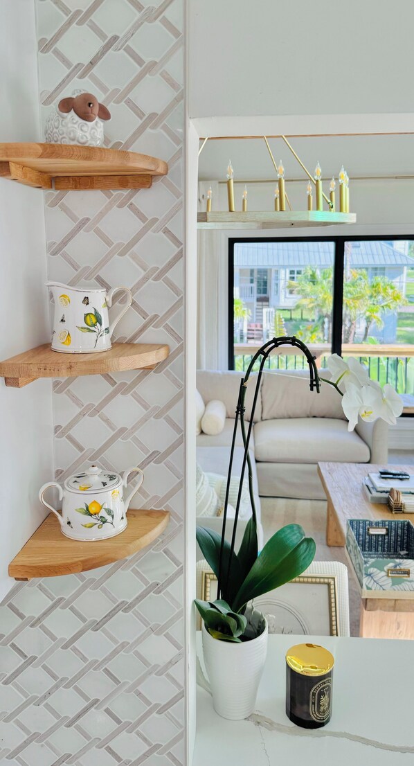 A living room with a sofa, coffee table, and decorative items. There are wooden shelves with kitchenware and a potted plant.