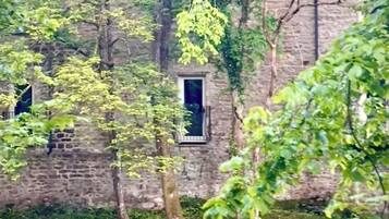 Ein rustikales Steinhaus mit kleinem Fenster, umgeben von üppiger Vegetation und einem Gewässer.