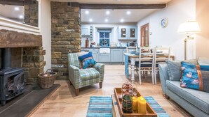 A cozy living room with a stone fireplace, a blue sofa, a plaid armchair, and a dining area with a table and chairs. - River Dance Cottage (Leyburn)