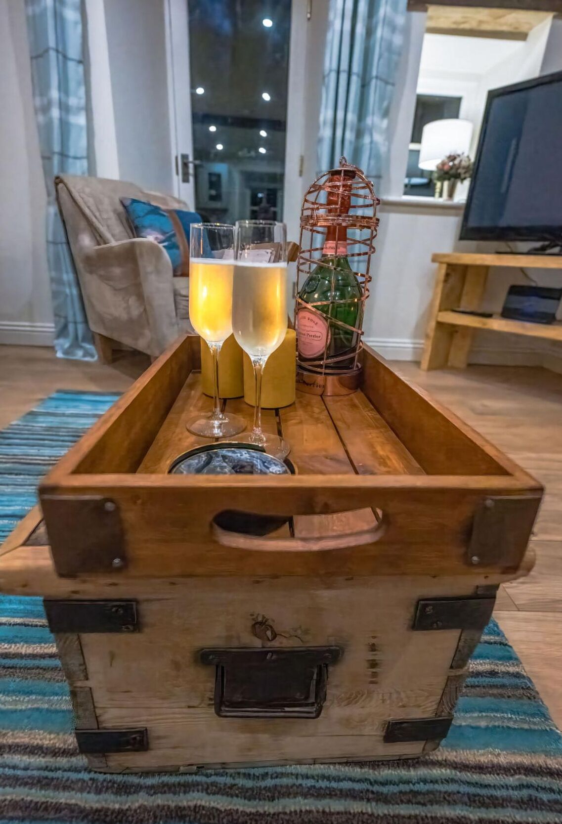 Een houten kist met twee champagneglazen en een fles wijn op een blauw-wit gestreepte vloerkleed.
