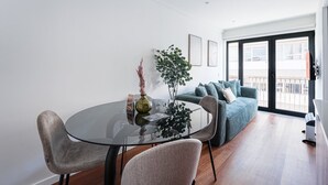 Ein modernes Wohnzimmer mit einem Glastisch, zwei Stühlen, einer Couch und einem großen Fenster mit Blick auf Gebäude.