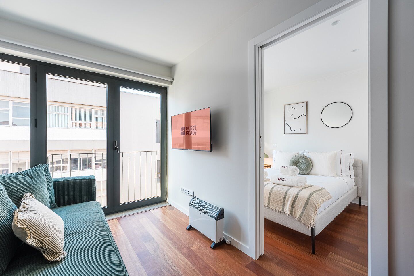 Een moderne hotelkamer met balkon, een bed met witte dekbedovertrek, een kleine verwarming en een tv aan de muur waarop staat: 'GUEST READY'.