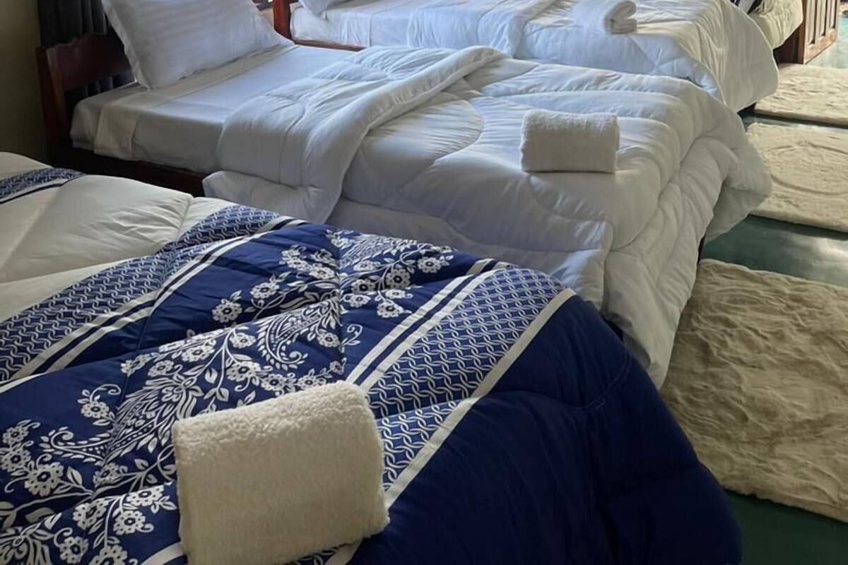 Une chambre d’hôtel avec trois lits, chacun recouvert de draps blancs et d’une couette à motifs floraux bleus. On y trouve des fenêtres avec des rideaux, un climatiseur fixé au mur et une porte en bois.