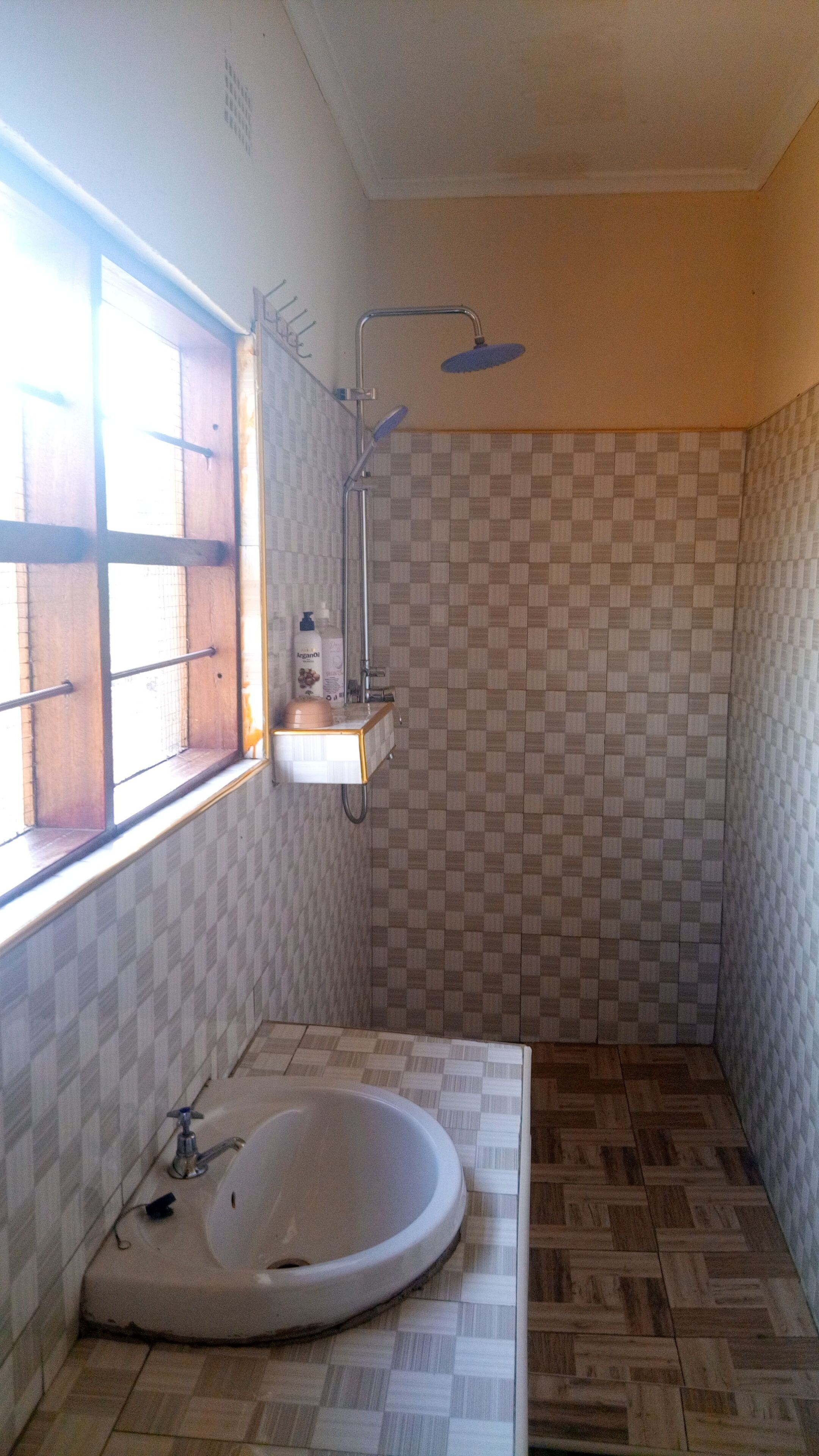 A bathroom with a checkered tile wall, a window with wooden blinds, a sink, and a shower with a handheld showerhead.