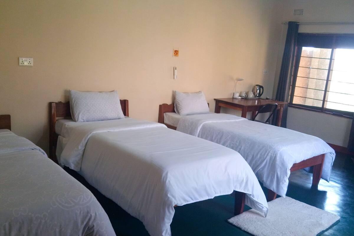 Une chambre d’hôtel avec trois lits simples, une petite table avec une bouilloire et une fenêtre donnant sur l’extérieur.