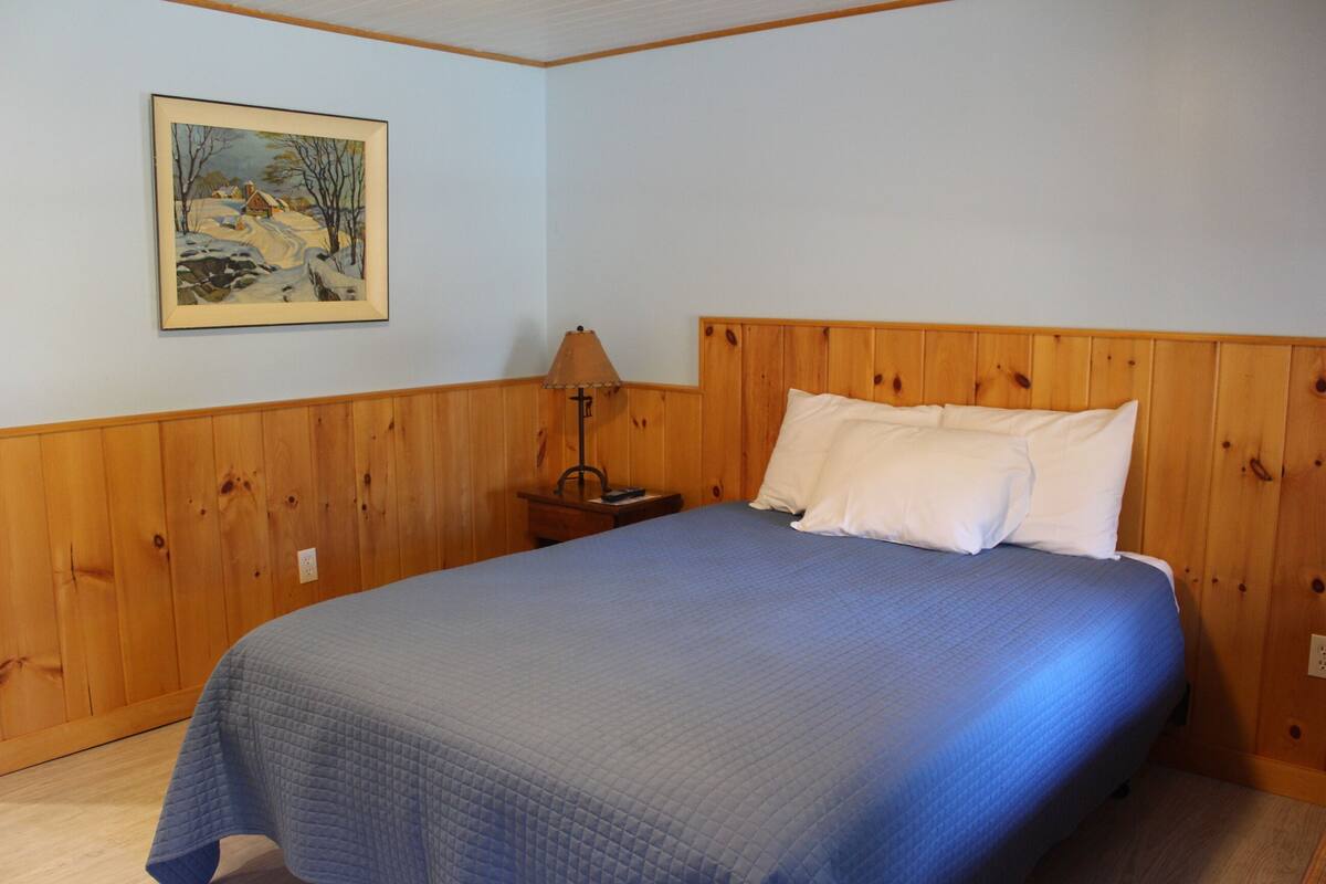 Quarto com cabeceira de madeira, colcha de cama matizada em azul, mesa de cabeceira com abajur e um quadro de uma paisagem nevada na parede.