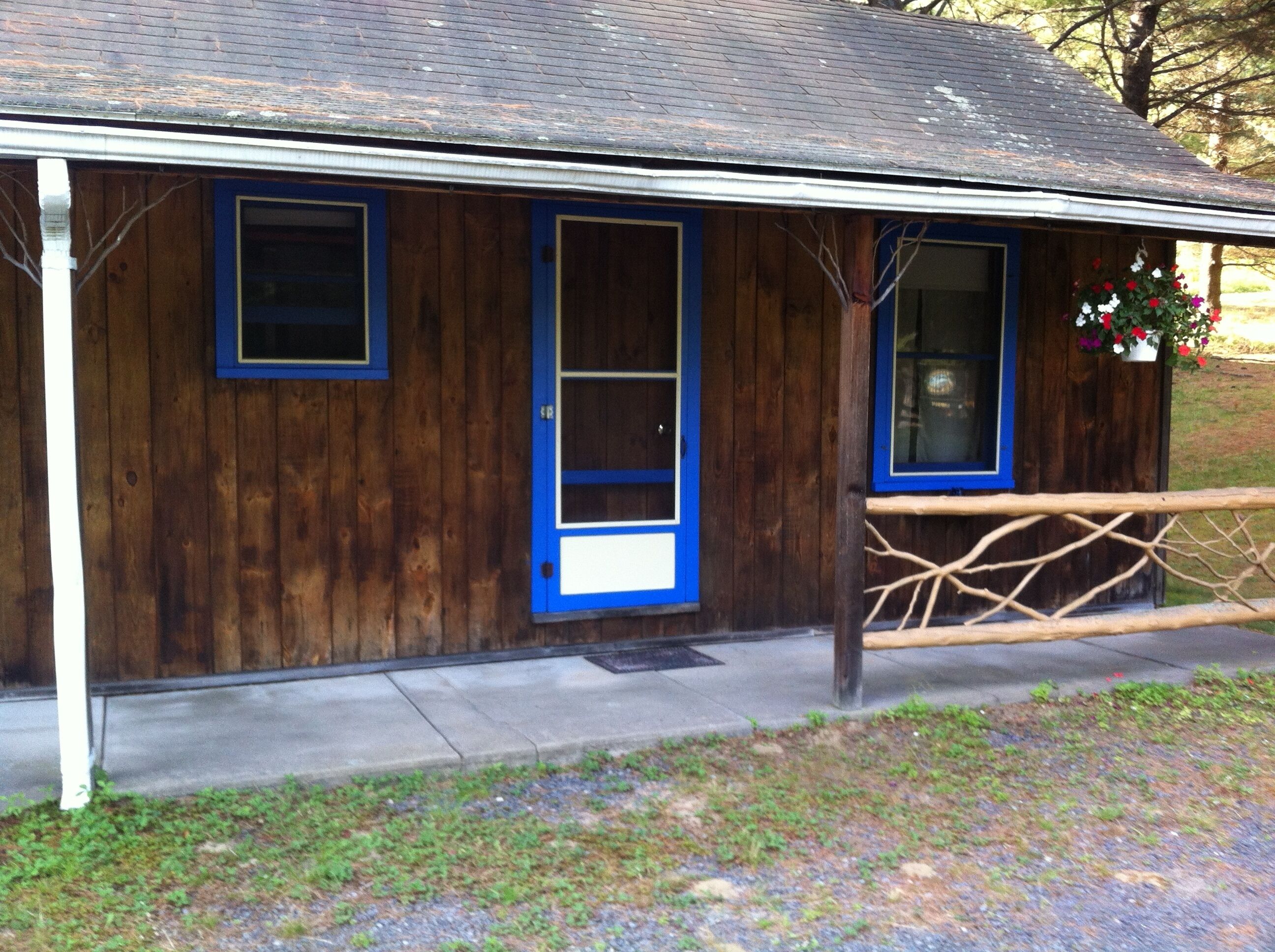 Un bâtiment rustique en bois, avec une porte et des fenêtres bleues, un porche doté d’une balustrade en bois et un petit jardin fleuri.