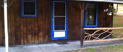 Un bâtiment rustique en bois, avec une porte et des fenêtres bleues, un porche doté d’une balustrade en bois et un petit jardin fleuri.