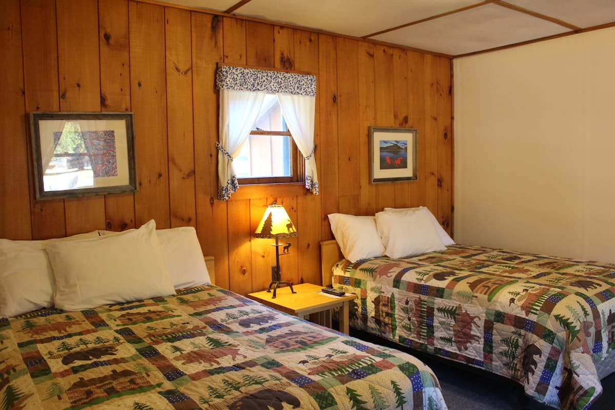 Quarto com duas camas, paredes de madeira, uma janela com cortina, uma luminária e quadros emoldurados.