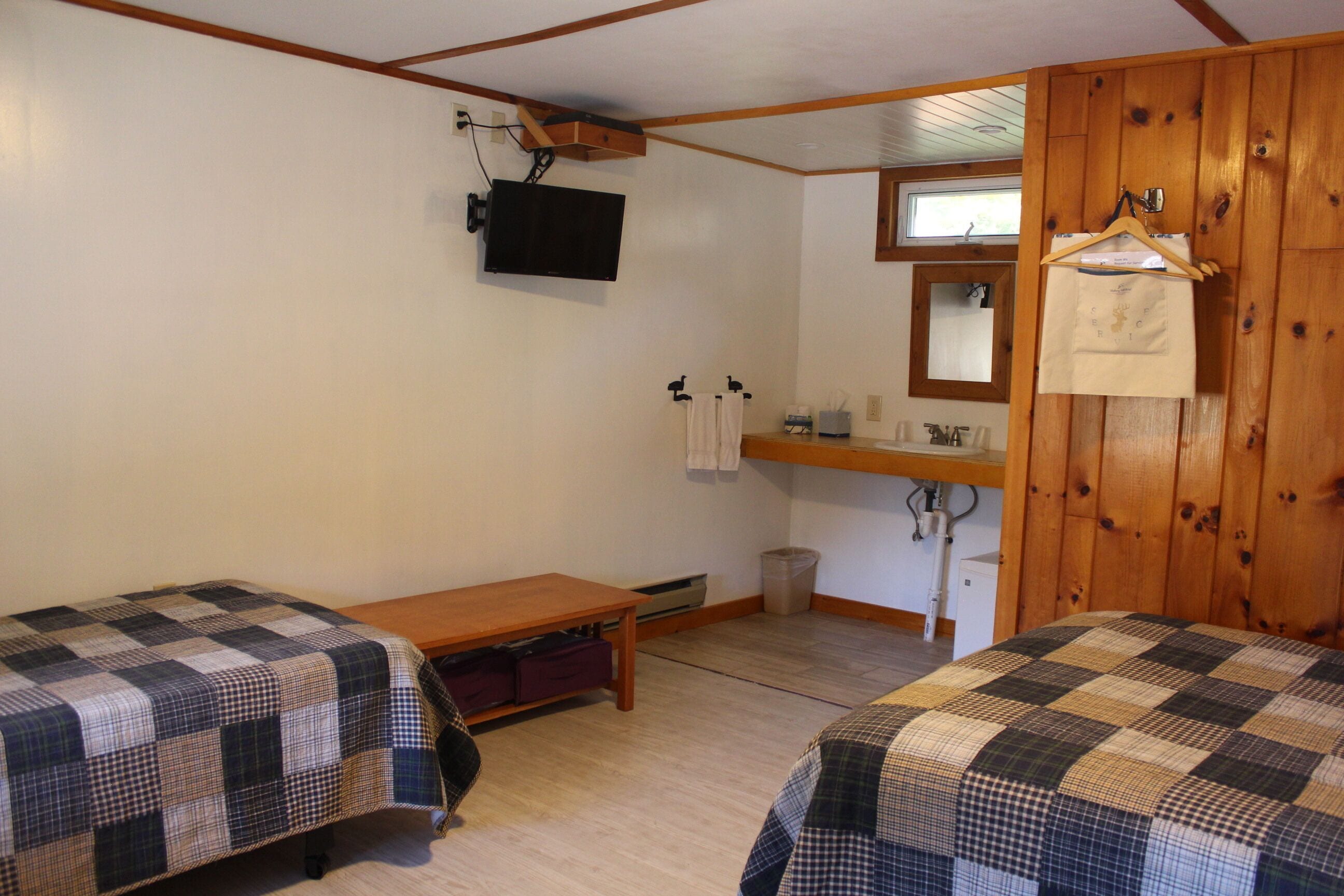 A room with two beds, a wooden wall, a television mounted on the wall, a small table, and a window.