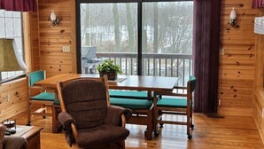 A wooden cabin interior with a dining table, chairs, and a large window with a view of trees.