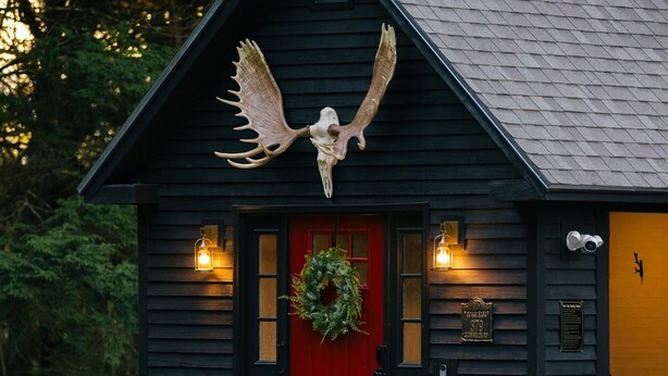 Un bâtiment rustique avec une porte rouge, une grande sculpture représentant une aile d’oiseau au-dessus de l’entrée et une guirlande sur la porte.