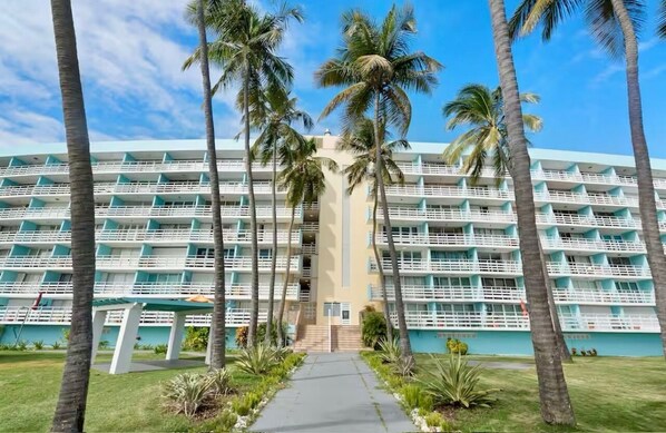 A multi-story hotel with a central staircase, surrounded by palm trees and a clear sky.