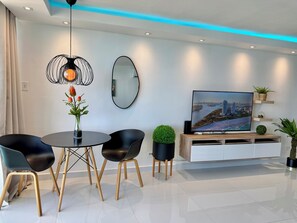 A modern living room with a round dining table, black chairs, a TV on a wooden stand, a mirror, and a cityscape on the screen. - Luxury Oceanfront Studio • Stunning Views • Pool • Balcony + Free Parking (Fajardo)