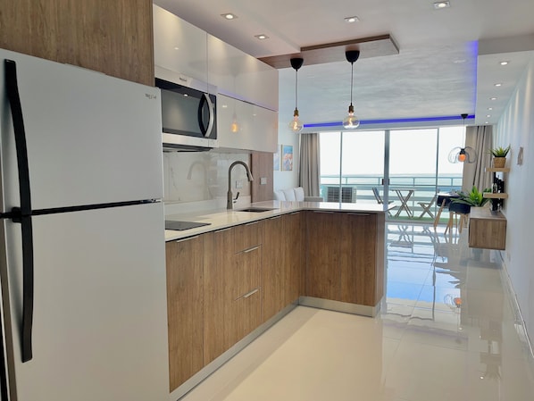 A modern kitchen with wooden cabinets, a stainless steel refrigerator, and a large window offering a view of the sea.
