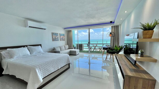 Quarto de hotel moderno com uma cama grande, área de estar com vista para o oceano e varanda com mobiliário exterior.