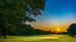 A golf course at sunset with a clear sky, trees, and a sand bunker.