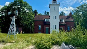 A lighthouse with a white tower and a windmill in a grassy area with trees.