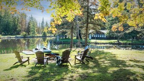 A lakeside seating area with Adirondack chairs and a fire pit, surrounded by trees and a dock with boats.