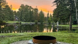 A serene lakeside setting with a metal fire pit surrounded by rocks, a wooden dock, and a small cabin in the background.